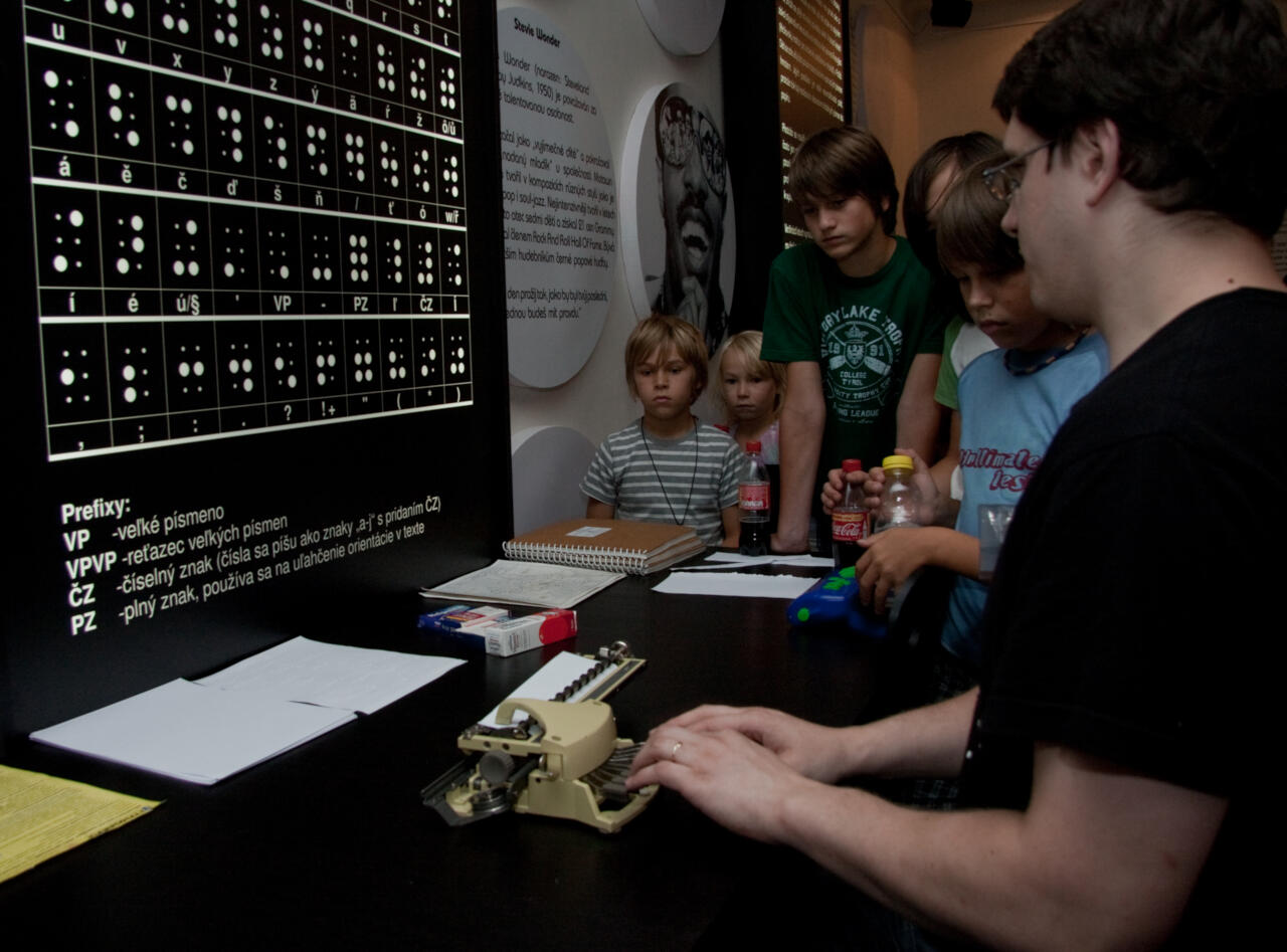 Muž píše Braillovým písmem u tabule na Neviditelné výstavě, obklopen dalšími návštěvníky